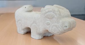 Escultura «Arkhos»: animal de cuatro patas tallado en piedra blanca, de formas estilizadas, con relieves en la cabeza y una cola enroscada, sobre una mesa