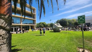Long queues of students beside a campus lawn during Welcome Day, with a large stone sculpture and palm trees