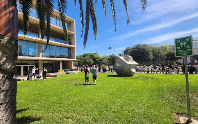 Long queues of students beside a campus lawn during Welcome Day, with a large stone sculpture and palm trees