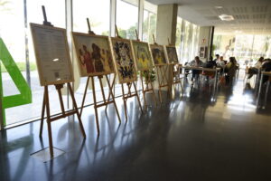 Caballetes de madera con las obras de la exposición Konngol Rewbe de la ONG AIDA y un panel informativo en el vestíbulo de la Casa del Estudiante