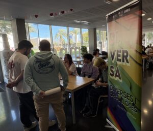 Mesa informativa del colectivo LGBTIQA+ DIVERSA, con su roll-up arcoíris y estudiantes conversando, bajo una guirnalda de corazones