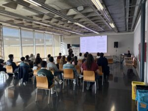 Assistents asseguts escolten un ponent al costat d'un roll-up de VRAIN i una gran pantalla a la sala Ximo Mora durant la jornada