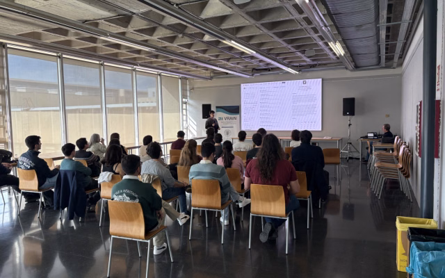 Assistents asseguts escolten un ponent al costat d'un roll-up de VRAIN i una gran pantalla a la sala Ximo Mora durant la jornada