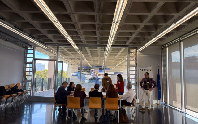 Participants working around a table during the European Visionary project session, beside its roll-up banner and the EU flag