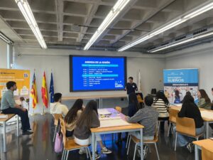 Un ponente presenta la agenda de la sesión a participantes sentados en mesas en la sala Ximo Mora, junto a pancartas de AIPC Pandora e Impulsando Sueños