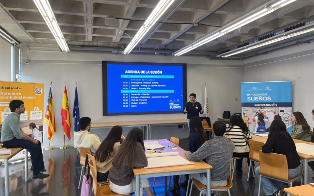 A speaker presents the session agenda to participants seated at tables in the Ximo Mora room, beside AIPC Pandora and Impulsando Sueños banners