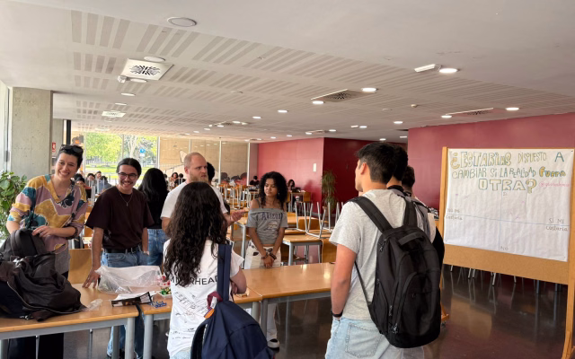 Estudiants conversen a la taula informativa del Grupo Bíblico Universitario, a la planta baixa de la Casa de l'Estudiant, al costat d'un panell d'enquesta