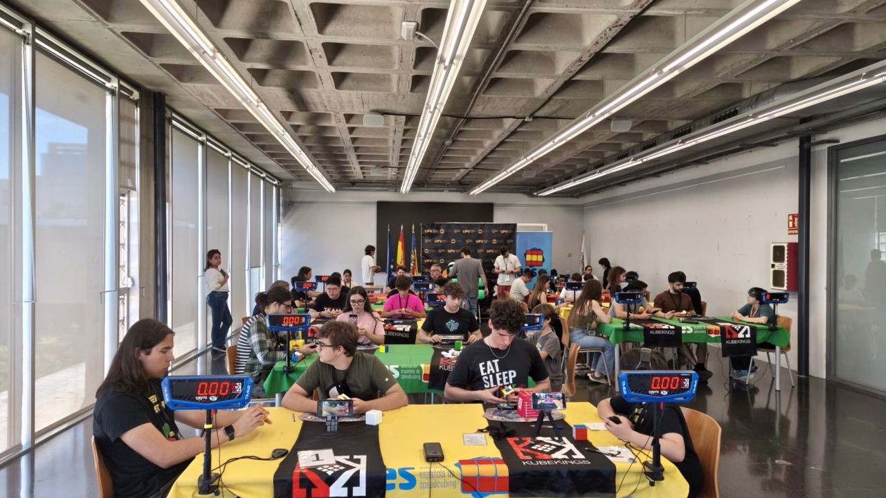 Participants resolen cubs de Rubik amb cronòmetres de speedcubing en taules amb tovalles grogues i verdes durant l'UPV Open 2026