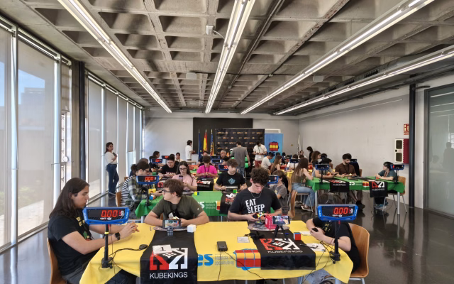 Competitors solve Rubik's cubes with speedcubing timers at tables with yellow and green tablecloths during the UPV Open 2026