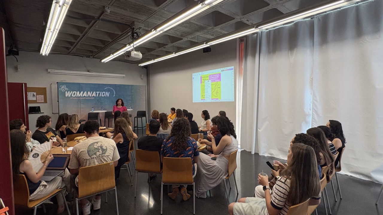 Públic assegut a les taules escoltant una ponència davant del fons de Womanation durant la jornada del Dia Internacional de la Dona en l'Enginyeria