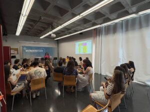 Públic assegut a les taules escoltant una ponència davant del fons de Womanation durant la jornada del Dia Internacional de la Dona en l'Enginyeria