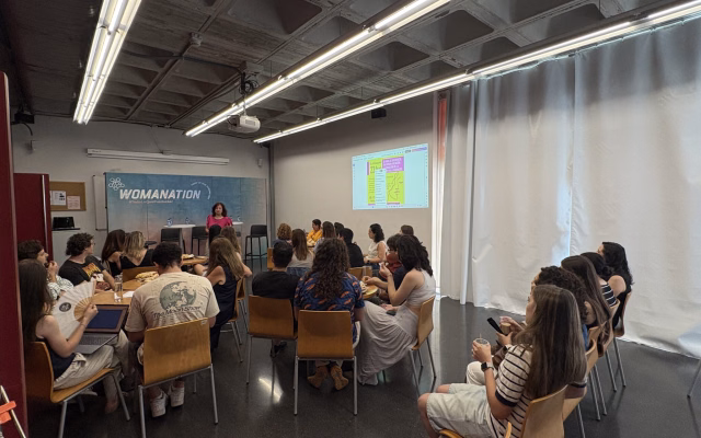 Audience seated at tables listening to a talk in front of a Womanation backdrop during the International Women in Engineering Day session