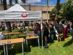 Students queue on the grass beside a Generación Espontánea tent with tables of drinks and leaflets during Welcome Day