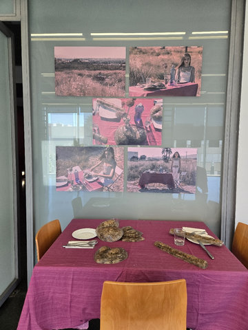 Instal·lació: taula amb tovalles fúcsia, plats i peces d'aspecte orgànic, davall de cinc fotografies rosades d'una dona i una taula al camp