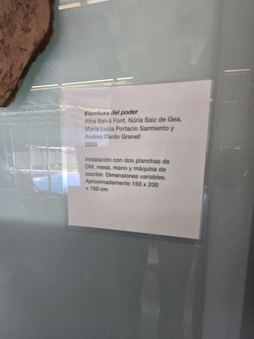 Cartel·la de l'obra «Escritura del poder» (2026): instal·lació amb dues planxes de DM, taula, mà i màquina d'escriure