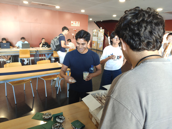 Un estudiant somrient agafa un got d'orxata i un dònut, amb estudiants treballant amb portàtils al fons de la planta baixa