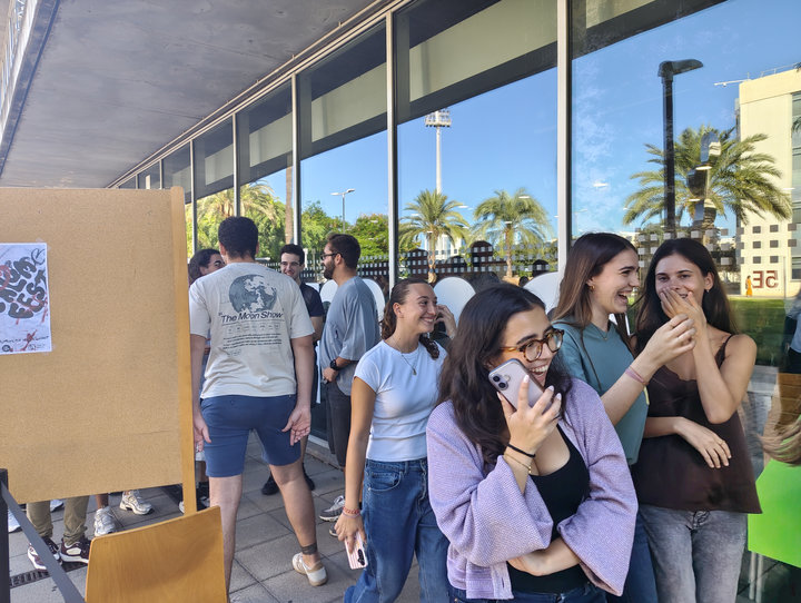 Estudiants somrients fan cua a l'exterior de la Casa de l'Estudiant, al costat d'un panell de suro amb un cartell