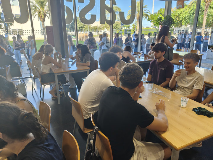 Estudiants asseguts a les taules de la planta baixa de la Casa de l'Estudiant amb gots d'orxata; darrere de la vidriera, més estudiants