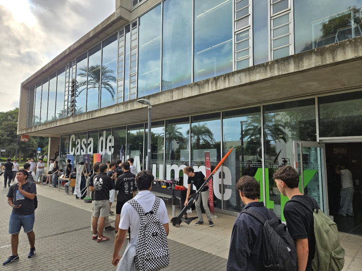 Un estudiante muestra un cohete naranja y negro sobre un soporte ante la fachada acristalada de la Casa del Estudiante, rodeado de estudiantes