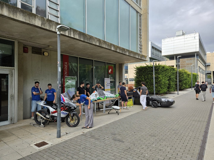 Moto de competición, prototipo sobre una mesa y coche aerodinámico negro expuestos por estudiantes en la acera junto a la Casa del Estudiante