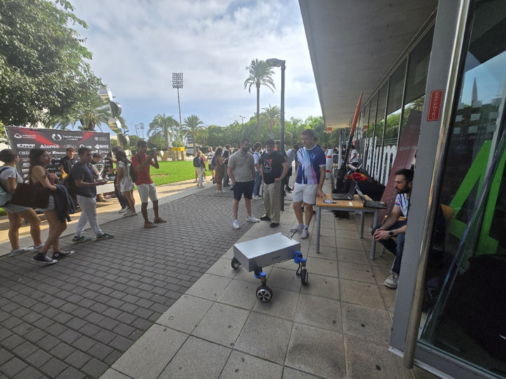 Estudiantes observan un pequeño robot de cuatro ruedas en la acera junto a la Casa del Estudiante, con césped y palmeras al fondo