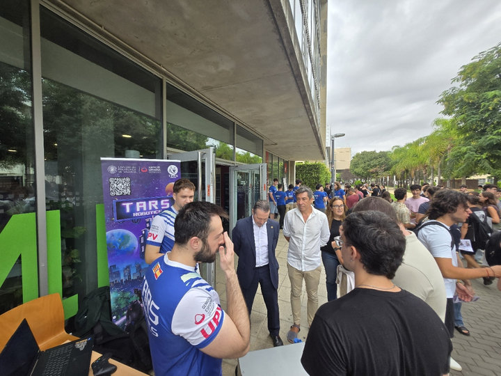 Miembros de TARS Robotics con camisetas de la UPV junto a su cartel enrollable, ante varios visitantes a la entrada de la Casa del Estudiante